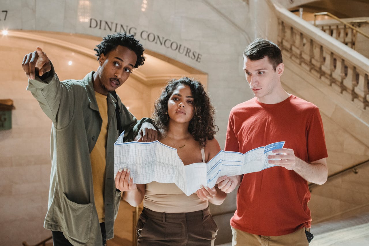 services-01 Young African American male tourist pointing away while searching for direction with diverse fiends standing in railway station terminal with paper map in hands