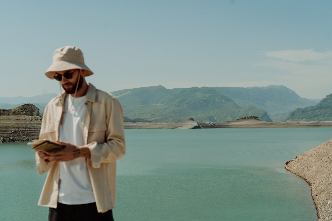 A man wearing a bucket hat and sunglasses looks at a book by a peaceful reservoir.